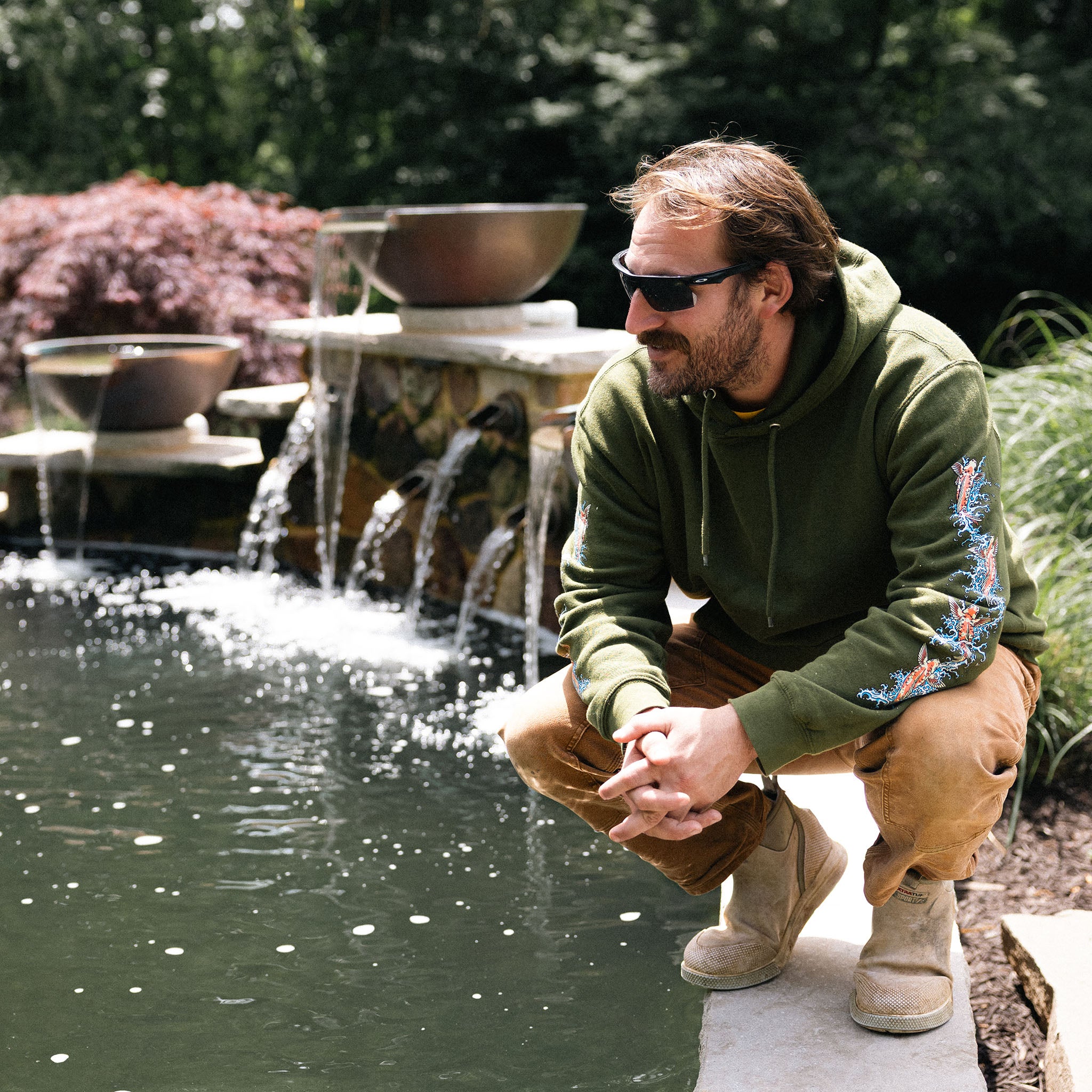 Cooper from Aqua Fauna Ponds in a green hoodie and sunglasses sitting by a water feature in a garden.